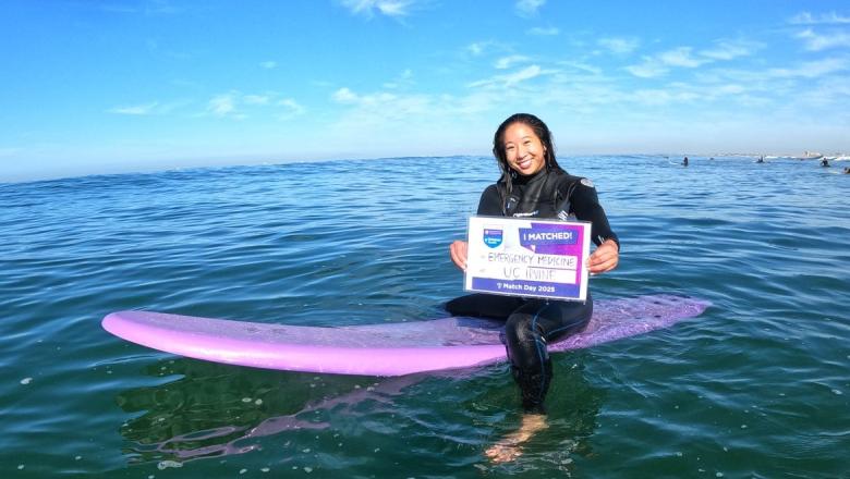 Cheyenne Low sitting on a surfboard in the ocean holding a card saying, 'I matched'